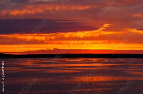 A fiery sunset is reflected in the calm river water  overhanging clouds in the bright orange sky  light waves on the water surface.