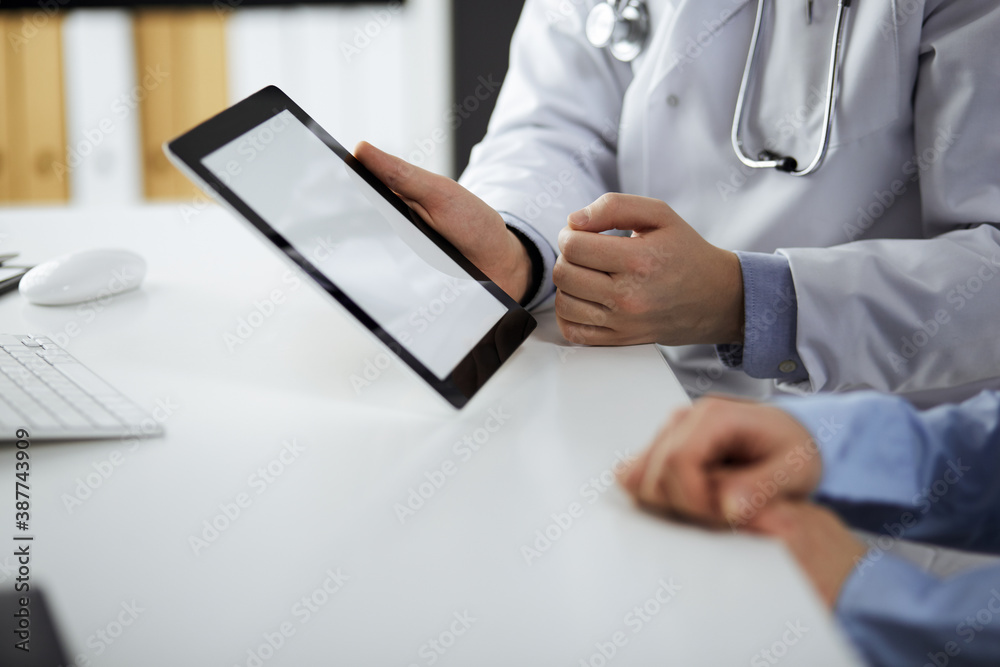 Unknown male doctor and patient woman discussing current health examination while sitting in clinic and using tablet computer, closeup of hands. Medicine and healthcare concept