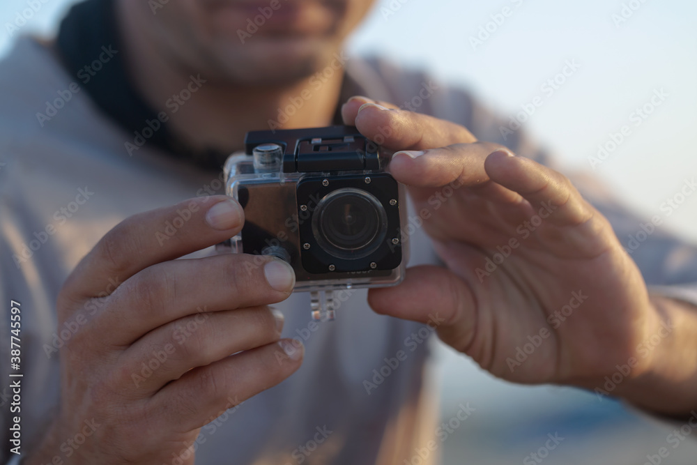 Small modern action camera in waterproof housing filming in shallow water on beach, man holding recording equipment sitting outdoors. Selective focus Small modern action camera in waterproof housing filming in shallow water on beach, man holding recording equipment sitting outdoors. Selective focus