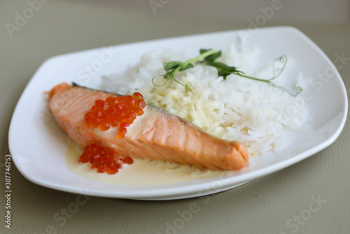 Grilled salmon steak, red caviar, boiled rice, milk sauce and peas micro green on whit plate on grey background
