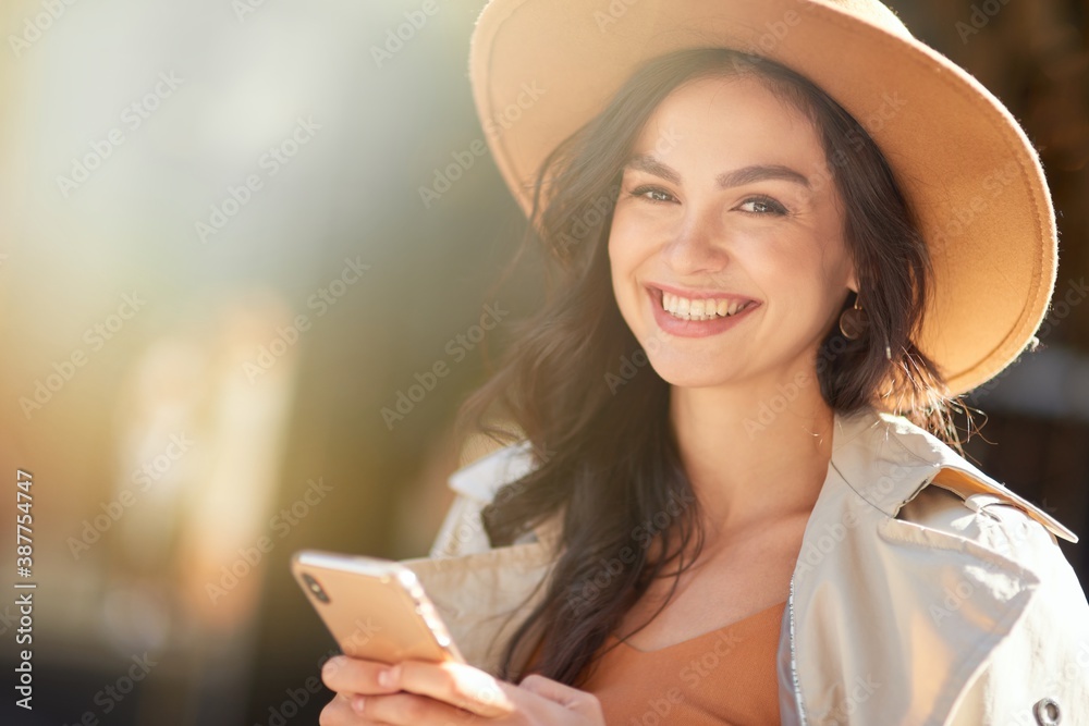 Portrait of a young beautiful and happy caucasian woman wearing hat using smartphone and smiling at camera while standing on the city street on a sunny day - obrazy, fototapety, plakaty 