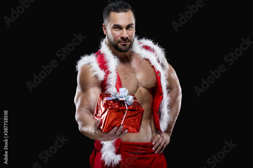 Muscular sexy sportsman in a red Santa Claus costume over naked body is holding out a Christmas gift isolated on black background