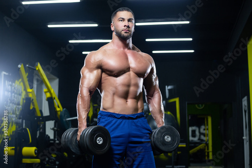 Front view of a muscular bodybuilder posing with dumbbells in blue shorts in a gym