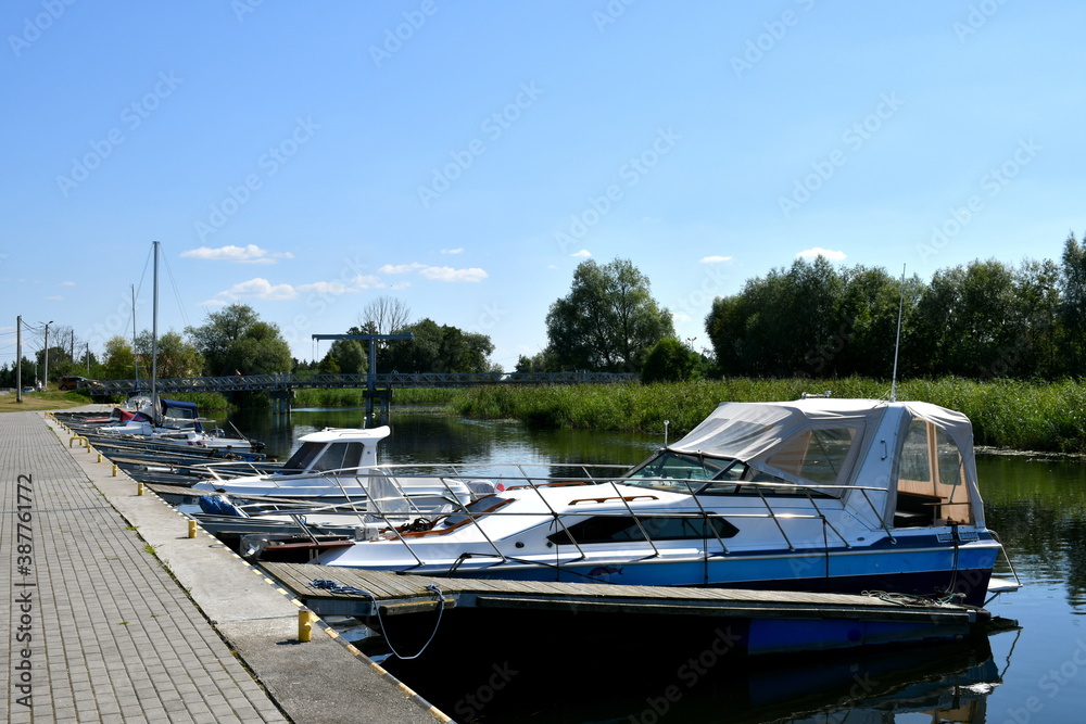 A view of a coastal concrete bank located next to a tiled pavement with a set of passenger boats, fishing vessels and other units parked next to it seen on a sunny summer day in Poland near the sea