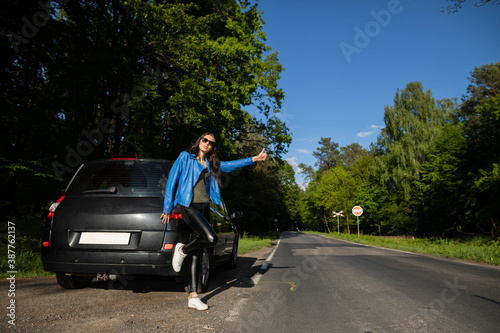 A young girl stands on the road in a green forest next to a broken car and asks for help, stopping other cars.