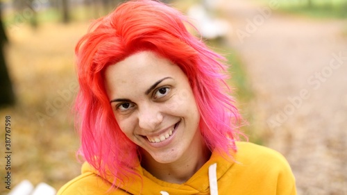 Portrait of a young pretty smiling woman with bright pink hair. Walk in the autumn park. Hipster attractive adult girl in oversized yellow hoodie or hoody. Positive emotions. Smile. 