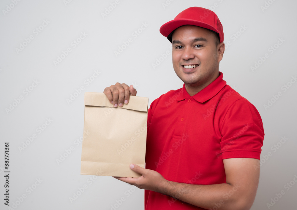 Young smiling asian delivery man in red uniform holding paper bag food delivery on isolated white background. Young smiling asian delivery man in red uniform holding paper bag food delivery on isolated white background.