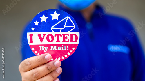 Man in mask from minority group produly holds Voted By Mail sticker during US Election closeup.