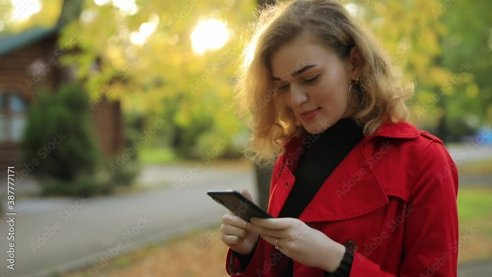 Beautiful woman looks down into smartphone at the fall day in a park and turn face at camera Beautiful woman looks down into smartphone at the fall day in a park and turn face at camera