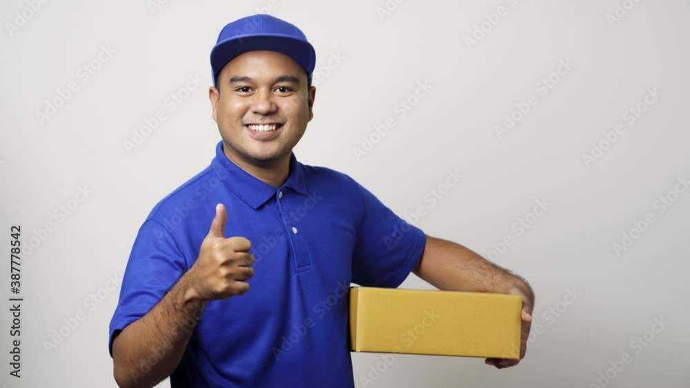Smiling young asian delivery man in blue uniform showing thumbs up holding parcel cardboard box on isolated white background. 4k Resolution.