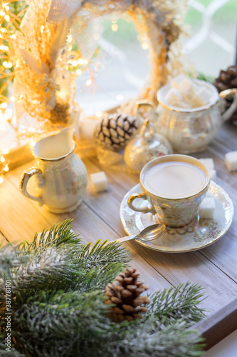 A cup of English tea with milk from an old mother-of-pearl porcelain service with refined sugar. Christmas tea party breakfast on the background of garlands and fir branches.