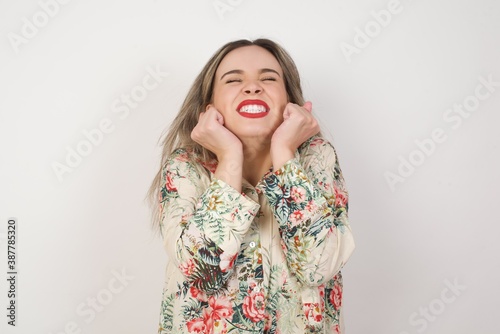 Portrait of young cute european student being overwhelmed with emotions, expressing excitement and happiness with closed eyes and hands near face while smiling broadly over gray background. © Jihan