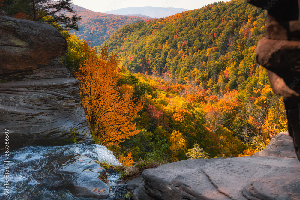 Water streaming off a rocky ledge leading into a valley valley fill with vibrant fall foliage color. Kaaterskill Falls - Woodstock, NY