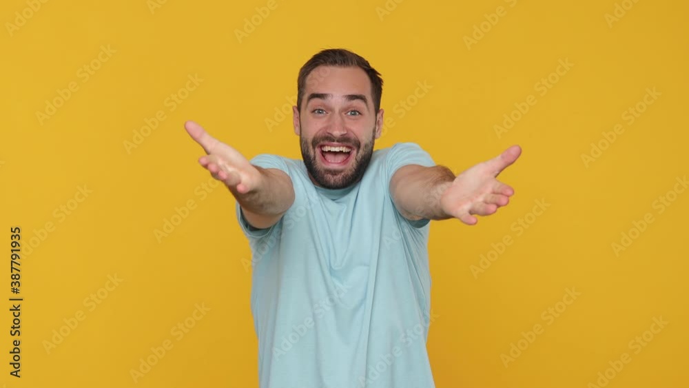 Bearded young man 20s in basic casual blue t-shirt looking camera charming smile standing with outstretched hands say come to me isolated on yellow background studio. People emotions lifestyle concept