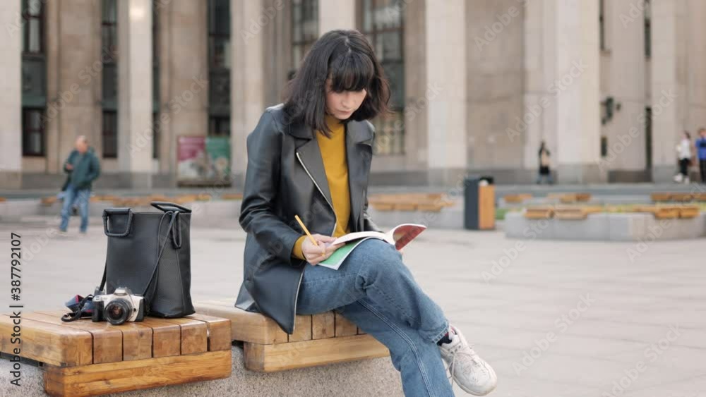 Young woman student working making notes outside in autumn. Preparing for classes near university, sitting on the bench with bag and photo camera Young woman student working making notes outside in autumn. Preparing for classes near university, sitting on the bench with bag and photo camera