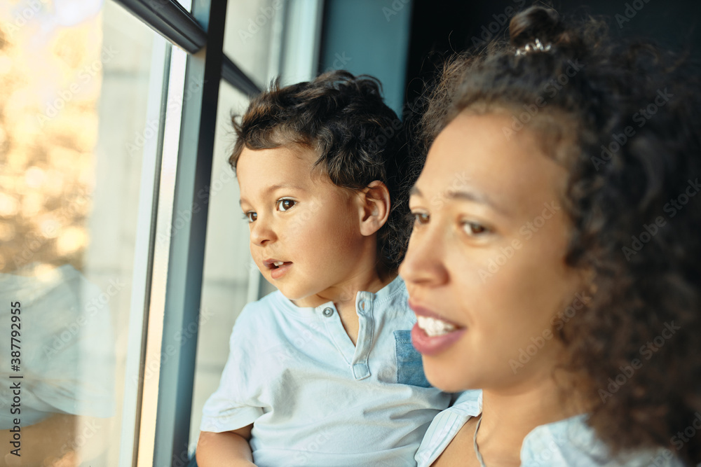 Family, togetherness, leisure and relaxation. Young mixed race female with little son in her arms having curious facial expression looking outside through window. Selective focus on boy face Family, togetherness, leisure and relaxation. Young mixed race female with little son in her arms having curious facial expression looking outside through window. Selective focus on boy face