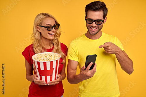 Funny young couple friends guy girl in 3d glasses isolated on yellow background. People in cinema concept. Watching movie film, hold bucket of popcorn, point index finger on mobile phone