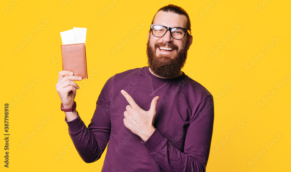 A young bearded man is holding a passport with some tickets in it and pointing at it is smiling at the camera . A young bearded man is holding a passport with some tickets in it and pointing at it is smiling at the camera .