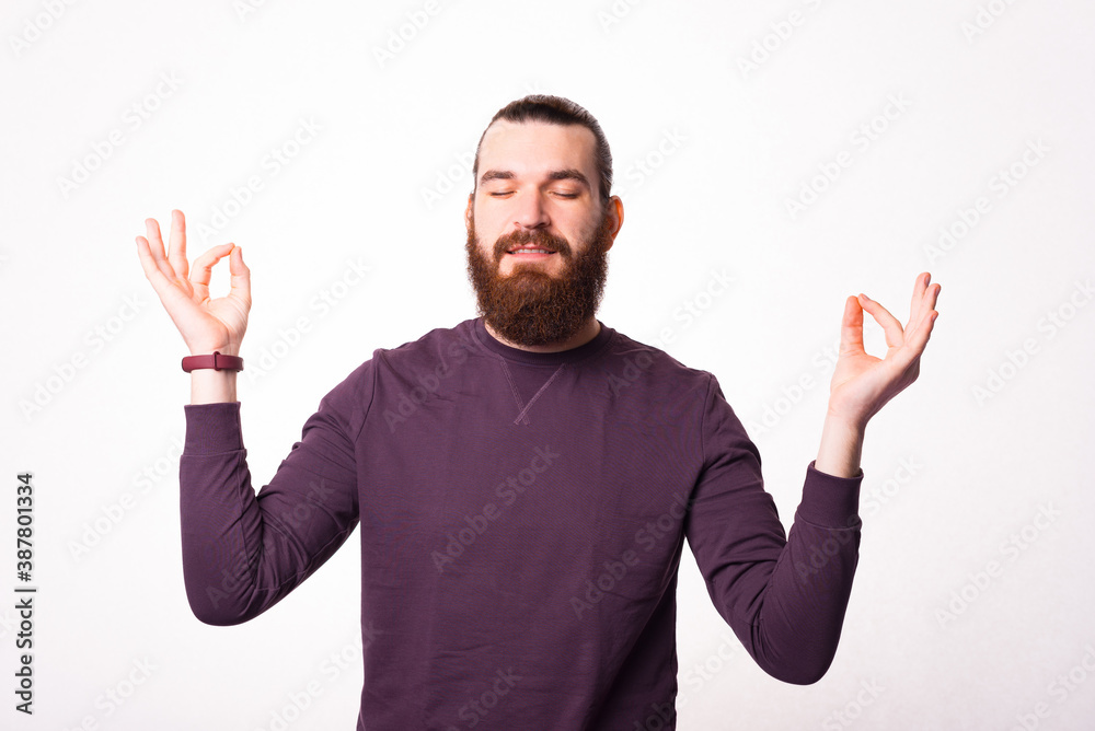 A young bearded man is sitting in relaxing pose near a white wall . A young bearded man is sitting in relaxing pose near a white wall .