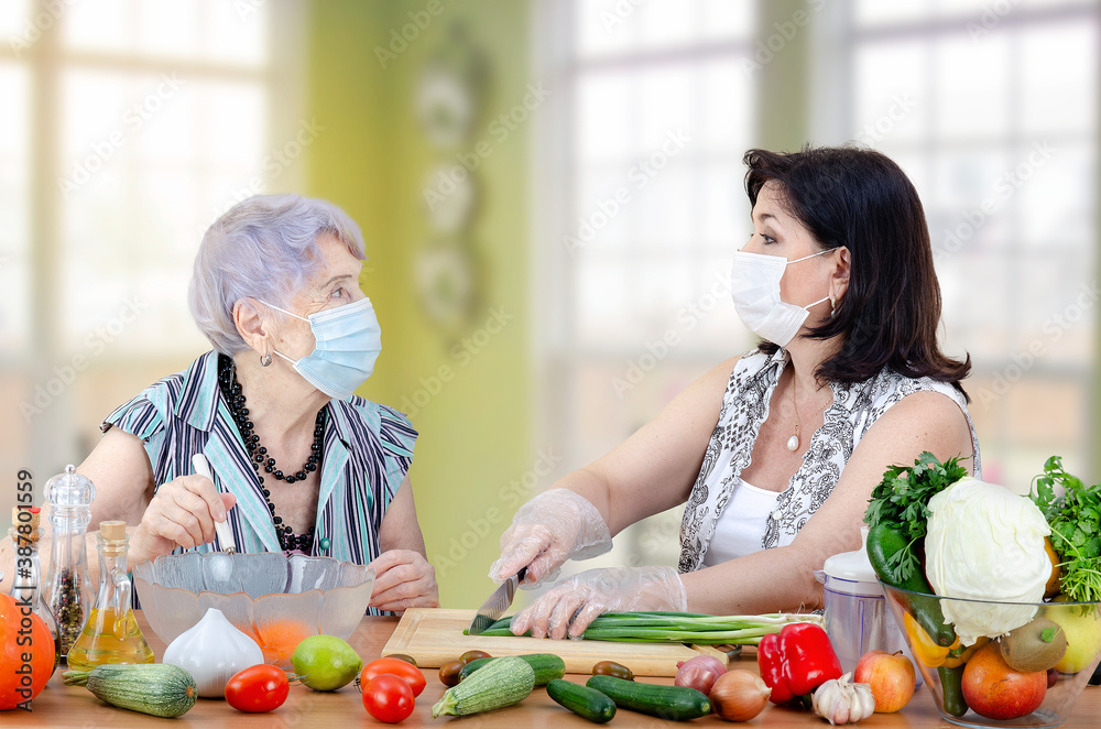Female caregiver and a senior adult woman are preparing a vegetable salad together. Both are wearing face protective masks due to the coronavirus pandemic. Female caregiver and a senior adult woman are preparing a vegetable salad together. Both are wearing face protective masks due to the coronavirus pandemic.