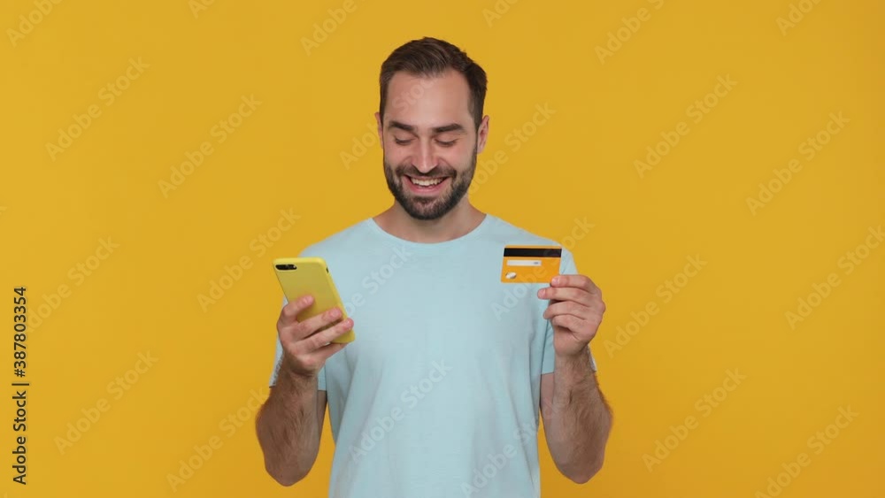 Happy fun excited young man in basic blue t-shirt using hold in hand credit bank card mobile cell phone just found out great big win news isolated on yellow background studio People lifestyle concept Happy fun excited young man in basic blue t-shirt using hold in hand credit bank card mobile cell phone just found out great big win news isolated on yellow background studio People lifestyle concept