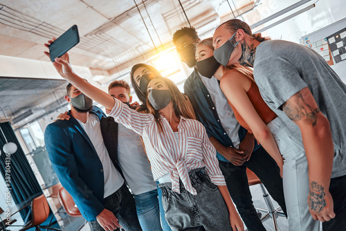 Young people with face masks back at work in office after lockdown, taking selfie.