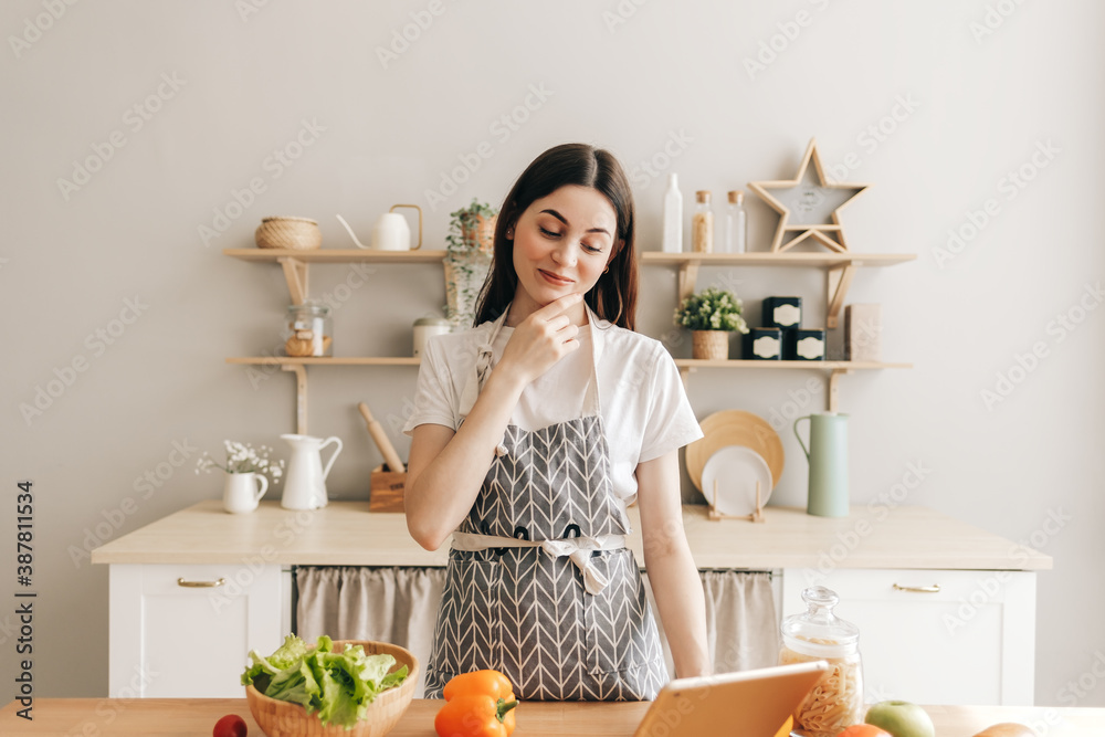 Young smiling caucasian woman in apron use tablet computer in the modern kitchen, preparing salad, read recipe.