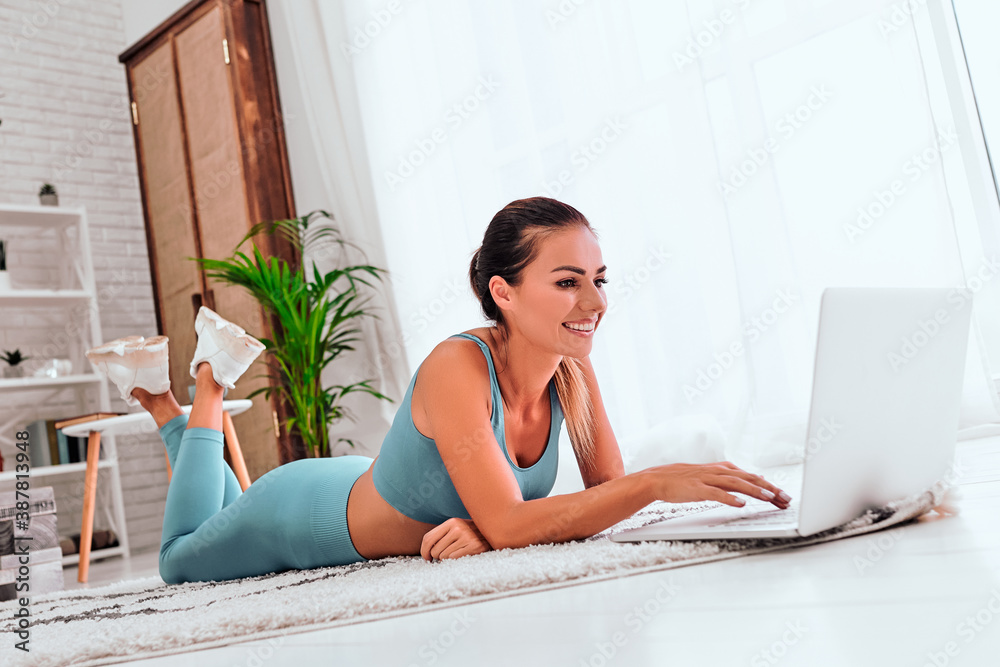 Glad, smiling and young woman in sportwear lying on floor and using laptop computer resting after training. Glad, smiling and young woman in sportwear lying on floor and using laptop computer resting after training.