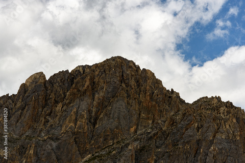 Close-up photo of a mountain peak in the French Alps, dark rock and clouds, blue sky. At the top very small mountaineers can be seen,