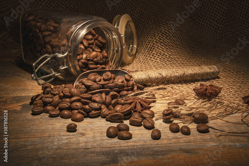 Vintage. A beam of warm light illuminates coffee beans casually scattered from a glass jar on a wooden table with burlap. Spoon for coffee beans.
