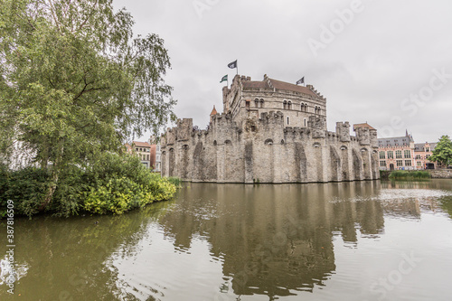 Panoramic view of calm waters surrounding the medieval castle of Gravensteen (Castle of the Counts), reflection in the water surface and green vegetation, cloudy day with a gray sky in Ghent, Belgium