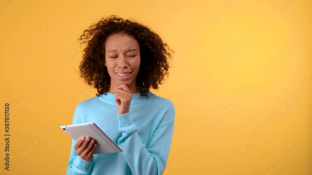puzzled young woman on a yellow background touches her chin and looks into the tablet. The woman makes a choice between several plaques. Isolated on a bright yellow background. 4K