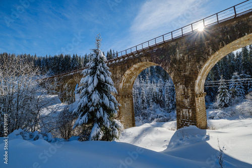 Stone viaduct (arch bridge) on railway through mountain snowy fir forest. Snow drifts on wayside and hoarfrost on trees and electric line wires.