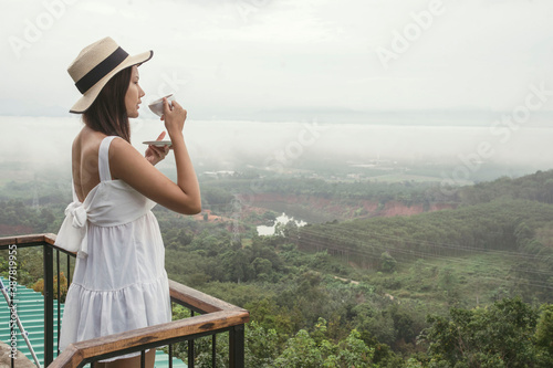 A beautiful Asian woman gets up in the morning drinking coffee on the top of the mountain, enjoying the view in the fog.
