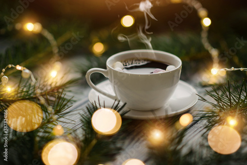A cup of hot coffee with marshmallows among the fir branches and lights of festive garlands.