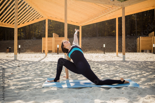 woman yoga in sports overalls performs Parivritta Parshvakonasana asana on a blue Mat on a sandy beach photo