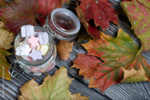 Marshmallows of different colors. They lie in a glass jar. Autumn maple leaves are scattered around.