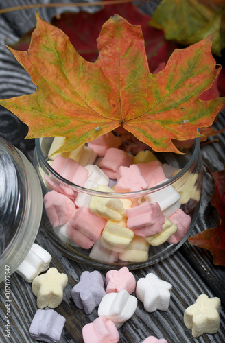 Marshmallows of different colors. They lie in a glass jar. Autumn maple leaves are scattered around.