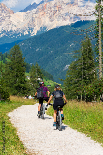 Group of mountain bikers riding their bicycles on a trail going through green meadows in the Italian Alps. Dolomite peaks are visible in the background. Val Badia, South Tyrol - Italy