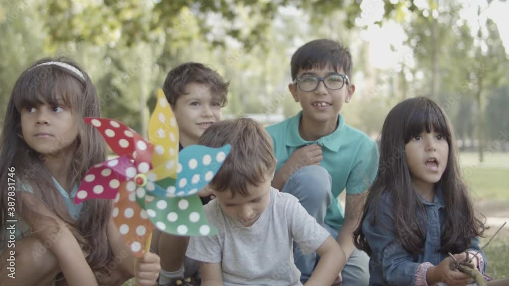 Group of multiethnic kids sitting together on open air lesson. Adorable children learning environment. Latin girl holding paper fan. Boy throwing ground. Childhood, education and summer school concept Group of multiethnic kids sitting together on open air lesson. Adorable children learning environment. Latin girl holding paper fan. Boy throwing ground. Childhood, education and summer school concept
