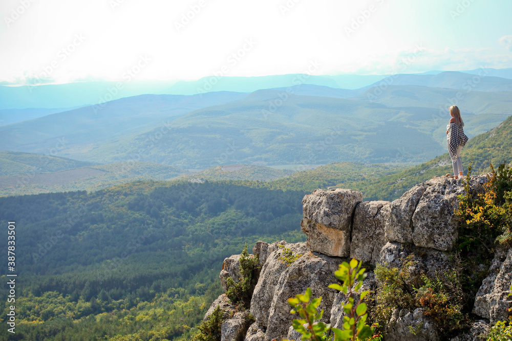 A young traveling woman stands on the edge of a mountain cliff and watching a beautiful view of the forest, mountains and clouds on vacation. The woman travels alone.