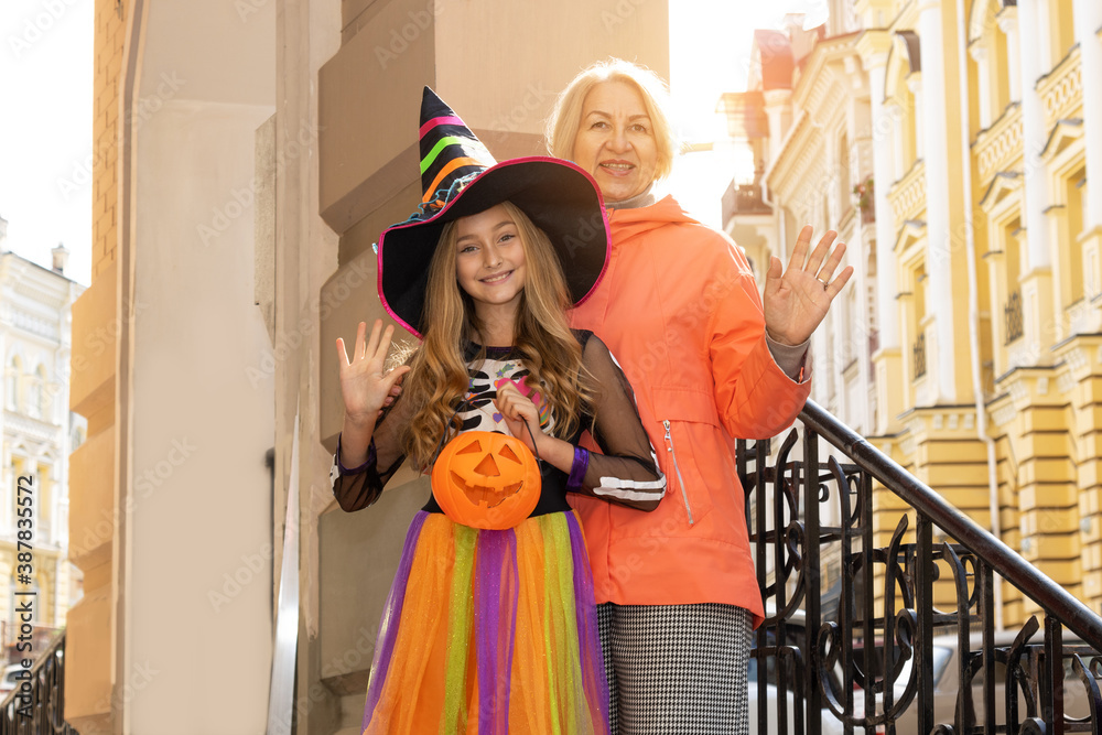 Happy Halloween! Grandmother and granddaughter. Girl in a witch costume holds jack pumpkin in her hands while standing with a mature woman outside her house.