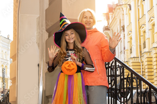 Happy Halloween! Grandmother and granddaughter. Girl in a witch costume holds jack pumpkin in her hands while standing with a mature woman outside her house. © A Stock Studio