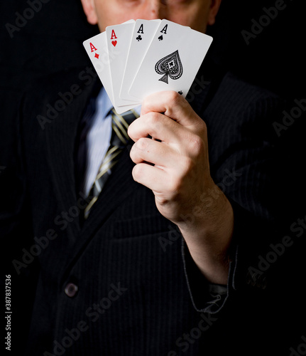 A man in a black suit and a tie holds 4 aces in his hand with black background. Poker cards. Play in poker and gambling games. Use for gambling sites, articles, advertising, gifts.