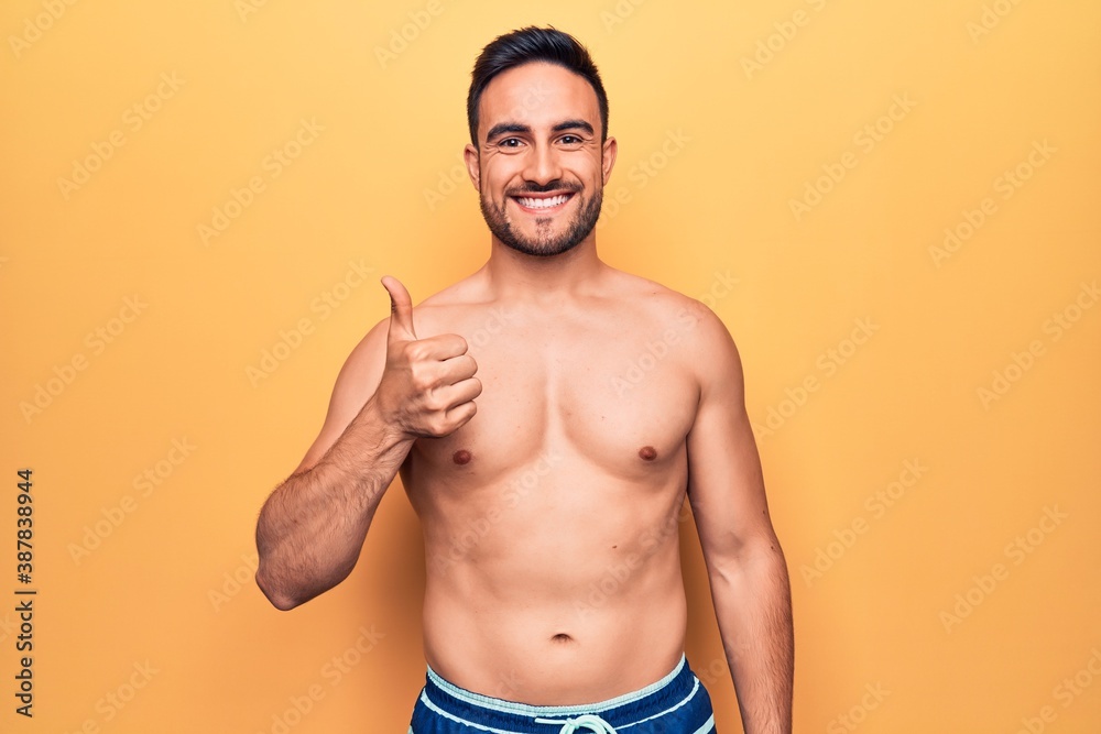 Young handsome man with beard wearing sleeveless t-shirt standing over yellow background Smiling happy and positive, thumb up doing excellent and approval sign