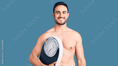 Young handsome man wearing swimwear holding weighing machine looking positive and happy standing and smiling with a confident smile showing teeth