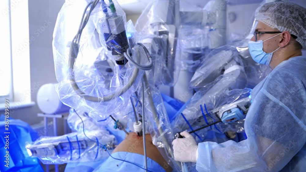 Microsurgery by medical robot. Doctor near the surgical arms of futuristic robotic machine performing a surgery to a patient. Hi-tech technology in medicine. Microsurgery by medical robot. Doctor near the surgical arms of futuristic robotic machine performing a surgery to a patient. Hi-tech technology in medicine.