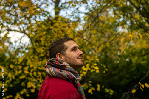 Photo of a young and attractive man wearing a scarf looking to the trees that surround him, enjoying a beautiful autumn day in nature