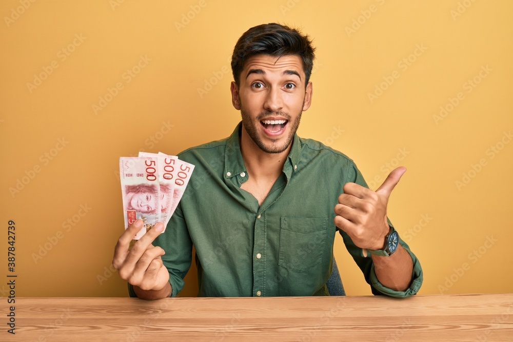 Young handsome man holding 500 swedish krona banknotes pointing thumb up to the side smiling happy with open mouth Young handsome man holding 500 swedish krona banknotes pointing thumb up to the side smiling happy with open mouth
