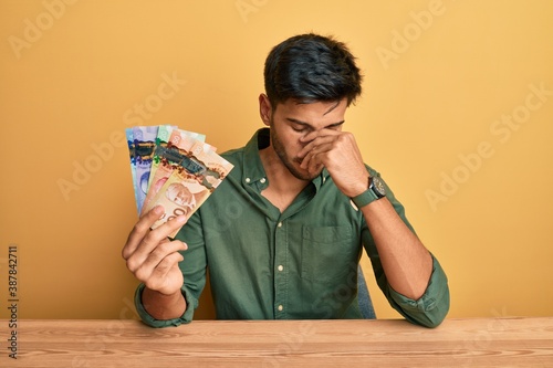 Young handsome man holding canadian dollars tired rubbing nose and eyes feeling fatigue and headache. stress and frustration concept.